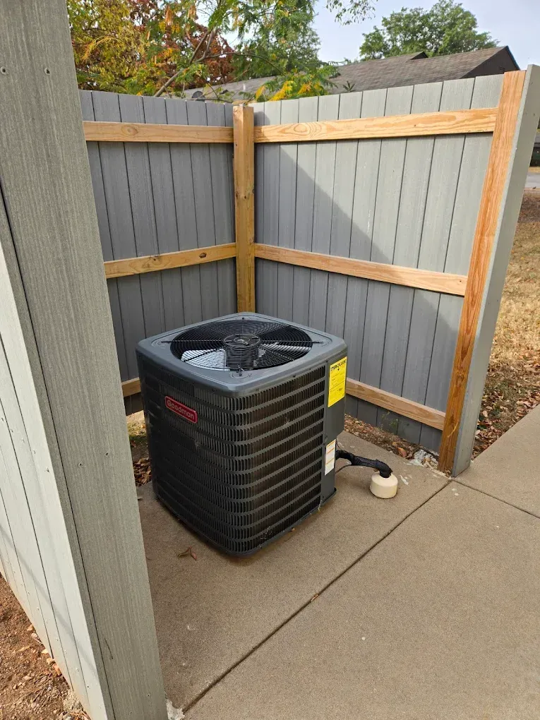 Outdoor air conditioner unit in a fenced corner beside a concrete patio, with gray and wood fencing.
