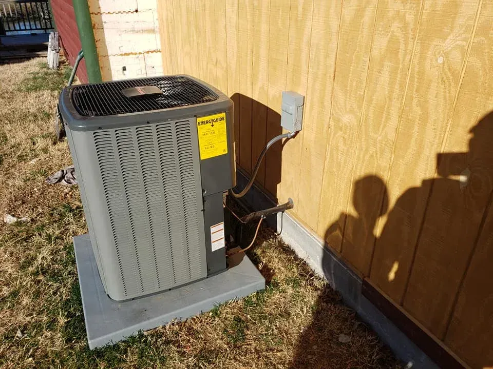 Outdoor air conditioner unit beside a wooden house wall on a concrete pad with electrical conduit nearby