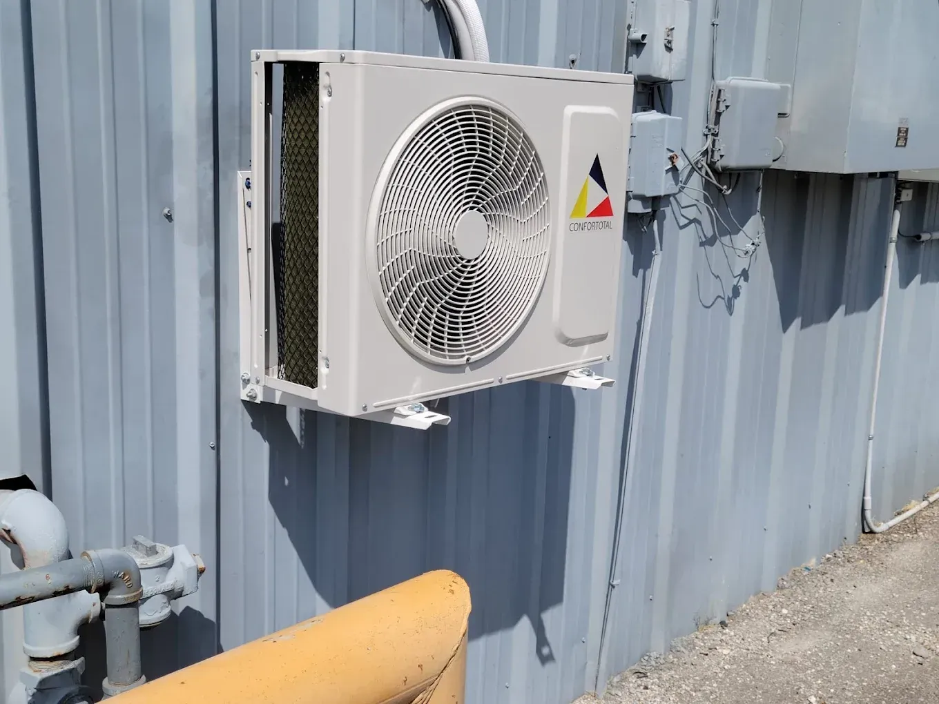White wall-mounted air conditioner unit on a blue corrugated metal building beside a yellow pipe and gravel ground