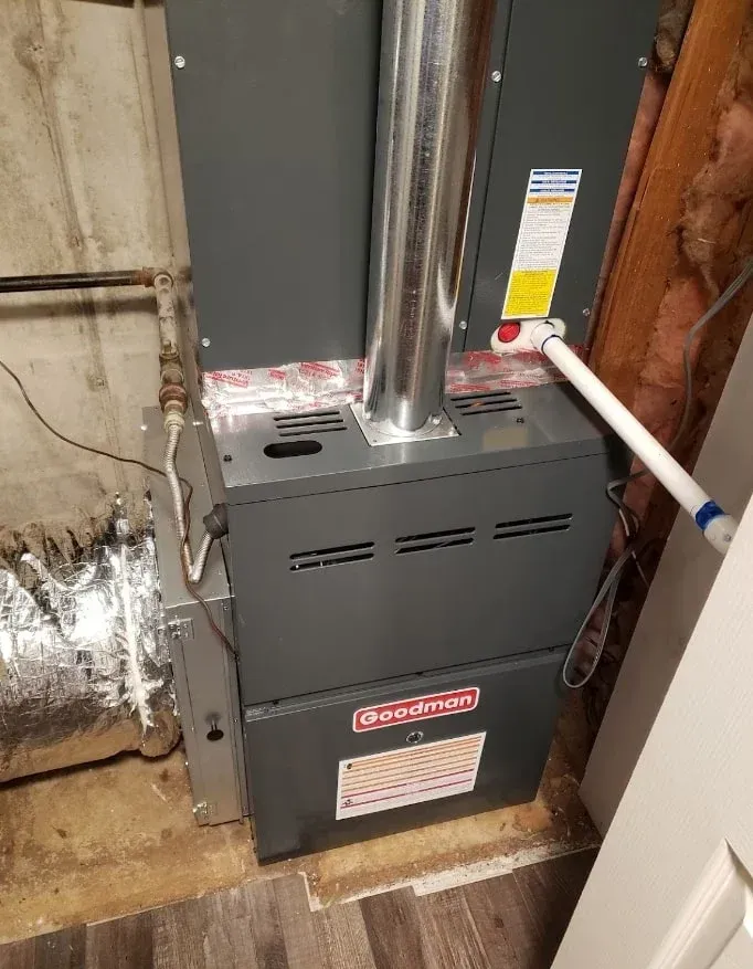 Basement furnace with metal vent pipes beside a white door and exposed insulation/wiring.