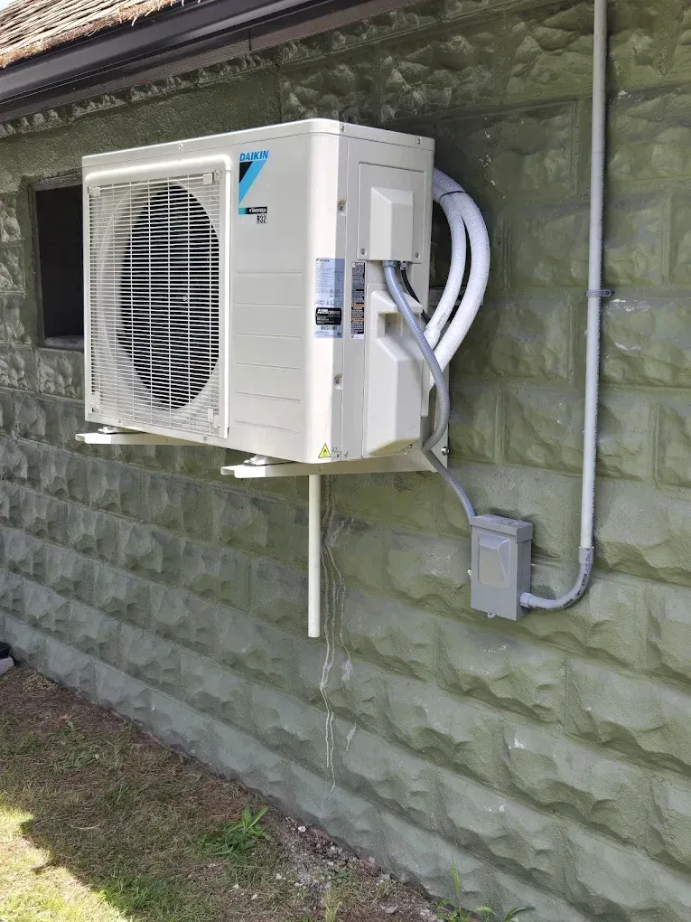 Wall-mounted outdoor air conditioner unit with exposed piping and conduit on a green exterior wall.