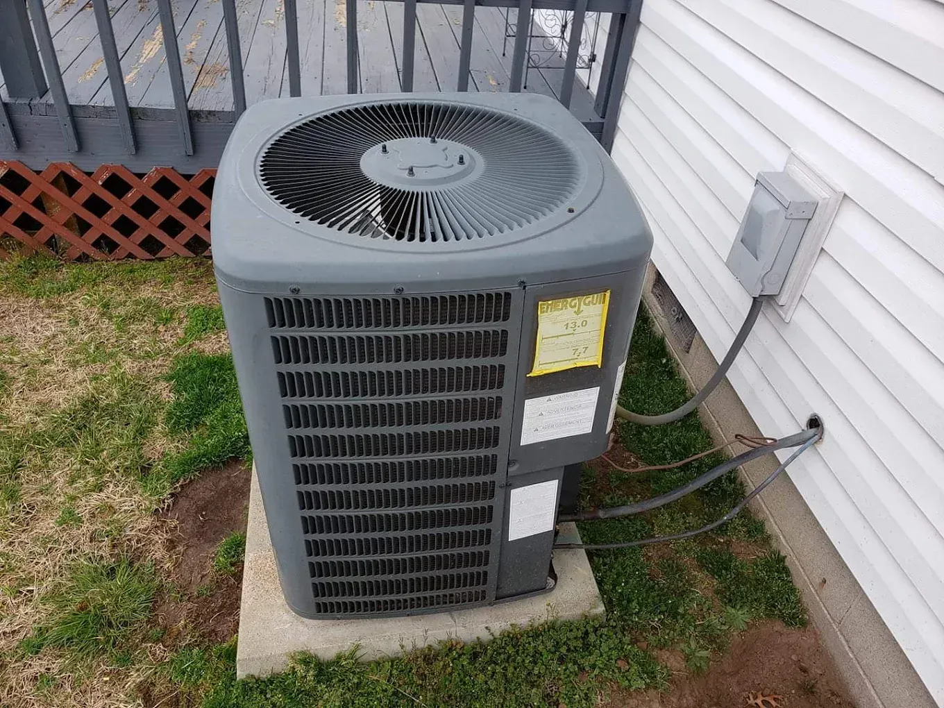 Outdoor central air conditioner unit beside a house wall and deck on a concrete pad