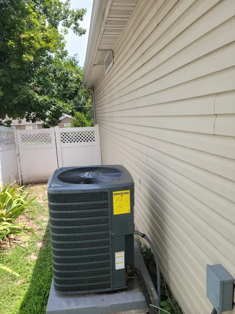 Outdoor air conditioner unit beside a beige house and white fence in a narrow side yard