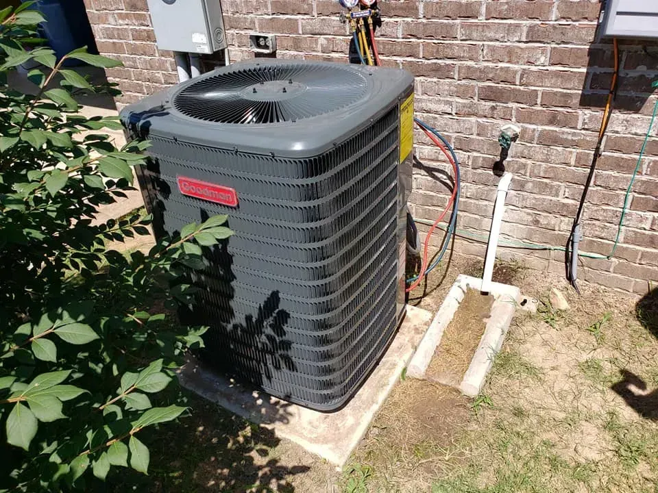 Outdoor HVAC unit beside a brick house with exposed pipes and wiring in a sunny yard