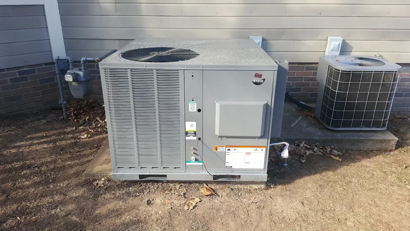 Outdoor HVAC unit beside a building wall, with a smaller condenser unit nearby on a dirt surface