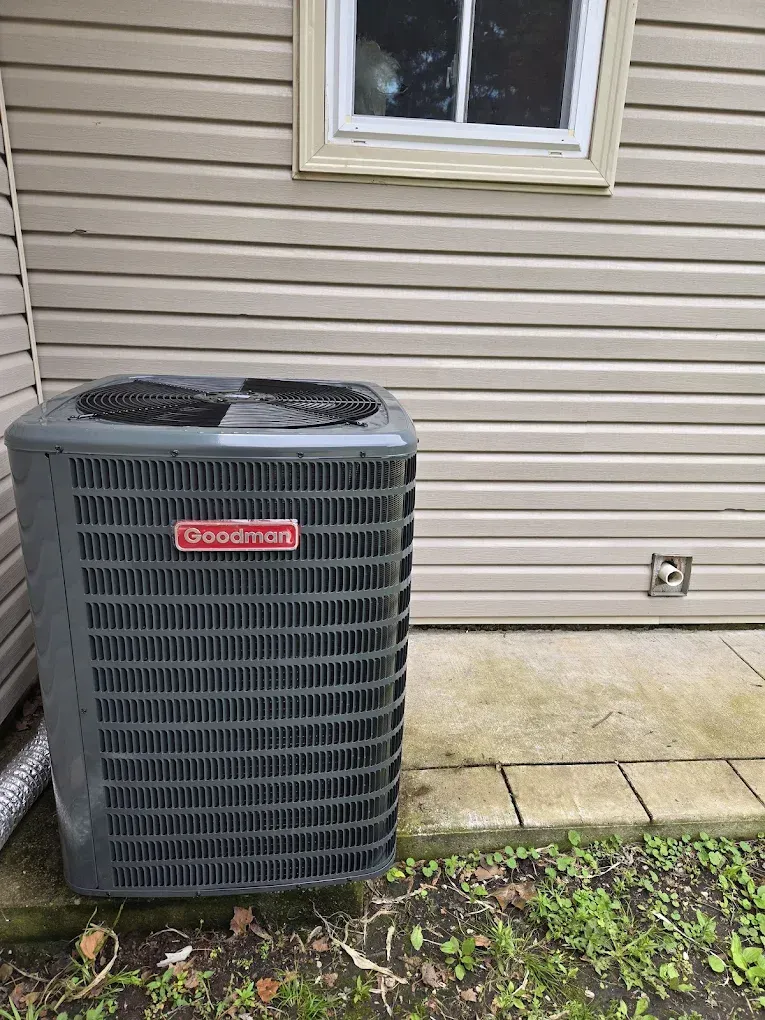 Outdoor Goodman air conditioner unit beside a house wall and concrete patio.
