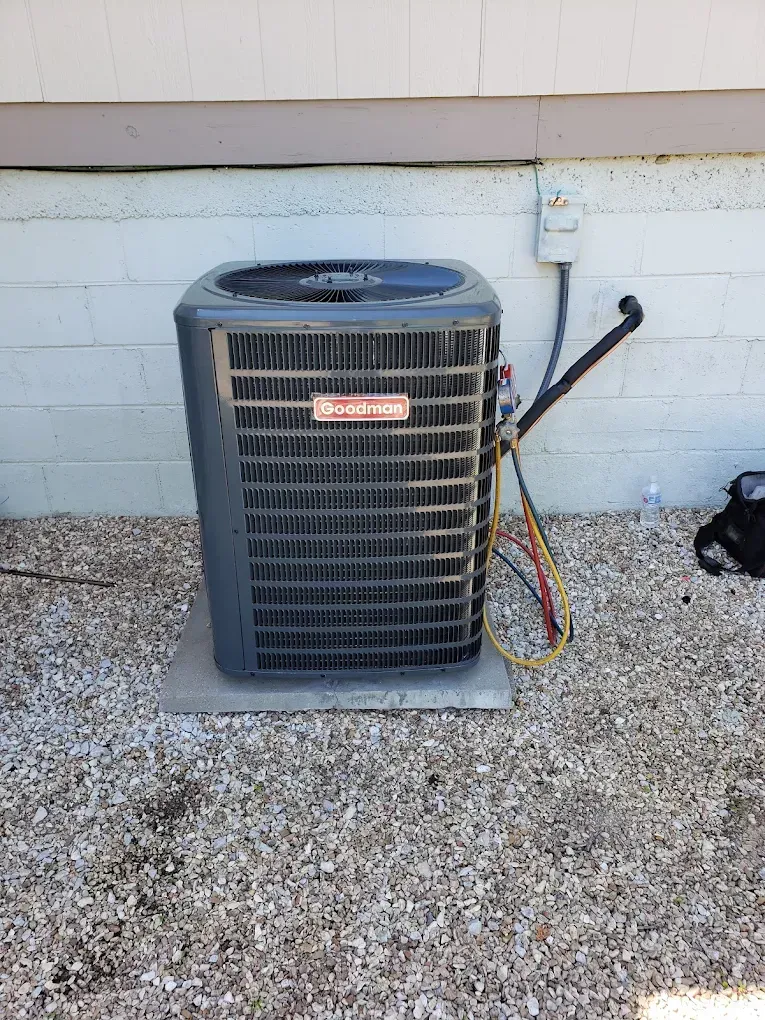 Outdoor HVAC condenser unit beside a house wall on a gravel patio, with exposed wires and hose nearby