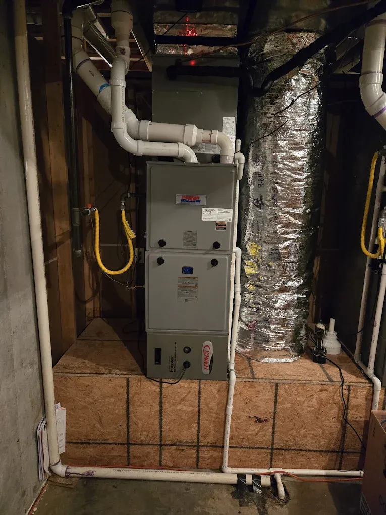 Basement utility area with a gray furnace, white pipes, foil ducting, and exposed brick wall.