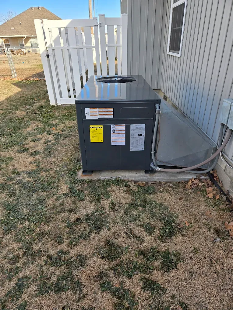 Outdoor HVAC unit beside a house, with a white fence and patchy grass in the yard.