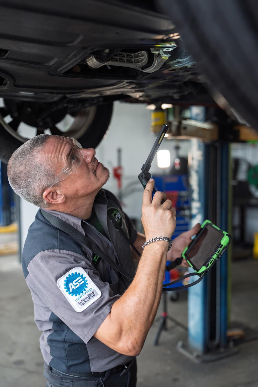 Mechanic inspecting a car undercarriage with a flashlight and diagnostic tablet in an auto shop