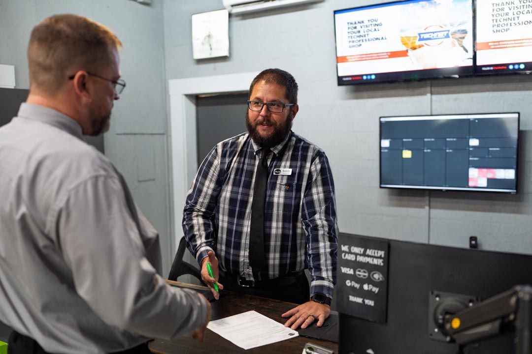 Two men talking at a counter in a control room with monitors on the wall.