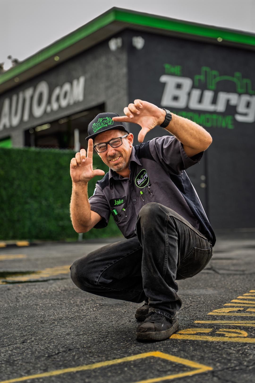 Smiling man crouches in front of a green-and-black building, posing with hands raised near the camera.