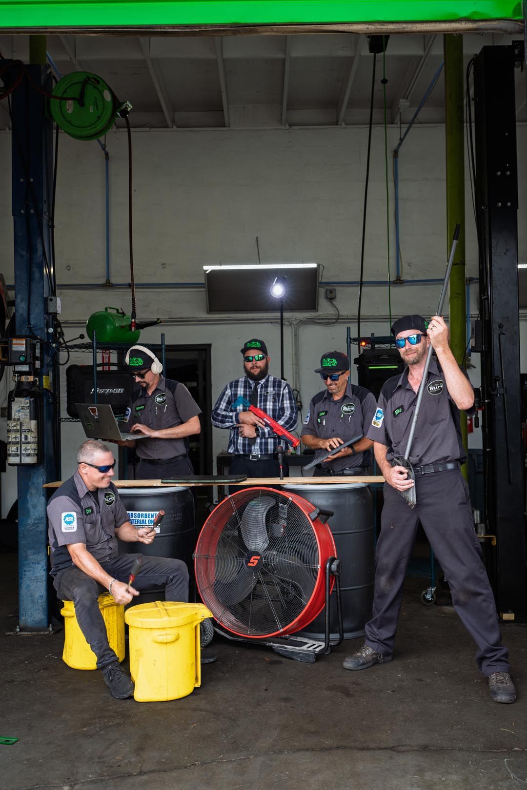 Five mechanics in a garage with a large red fan and yellow bucket, one kneeling by a car lift.