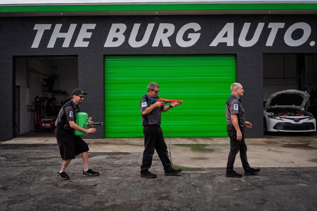 Three people in black shirts pose with colored boxes outside The Burg Auto garage with a bright green door.