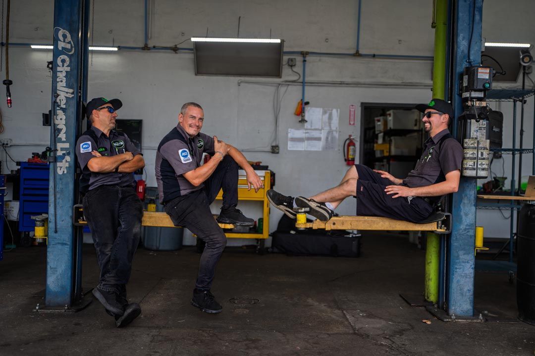 Three mechanics in dark uniforms posing in an auto garage around a yellow car lift
