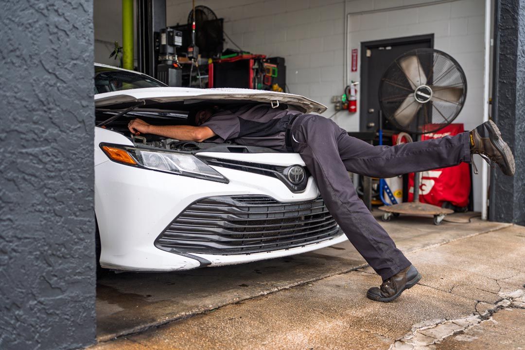 Person with legs sticking out from under a white car in a garage, near a large fan.