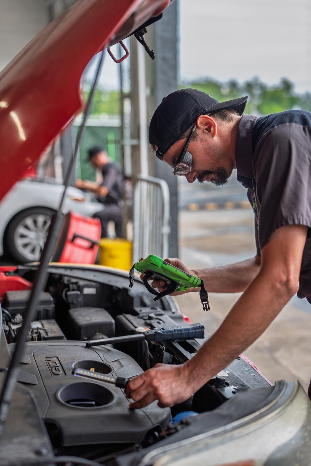 Mechanic working under an open car hood with a green tool in a garage