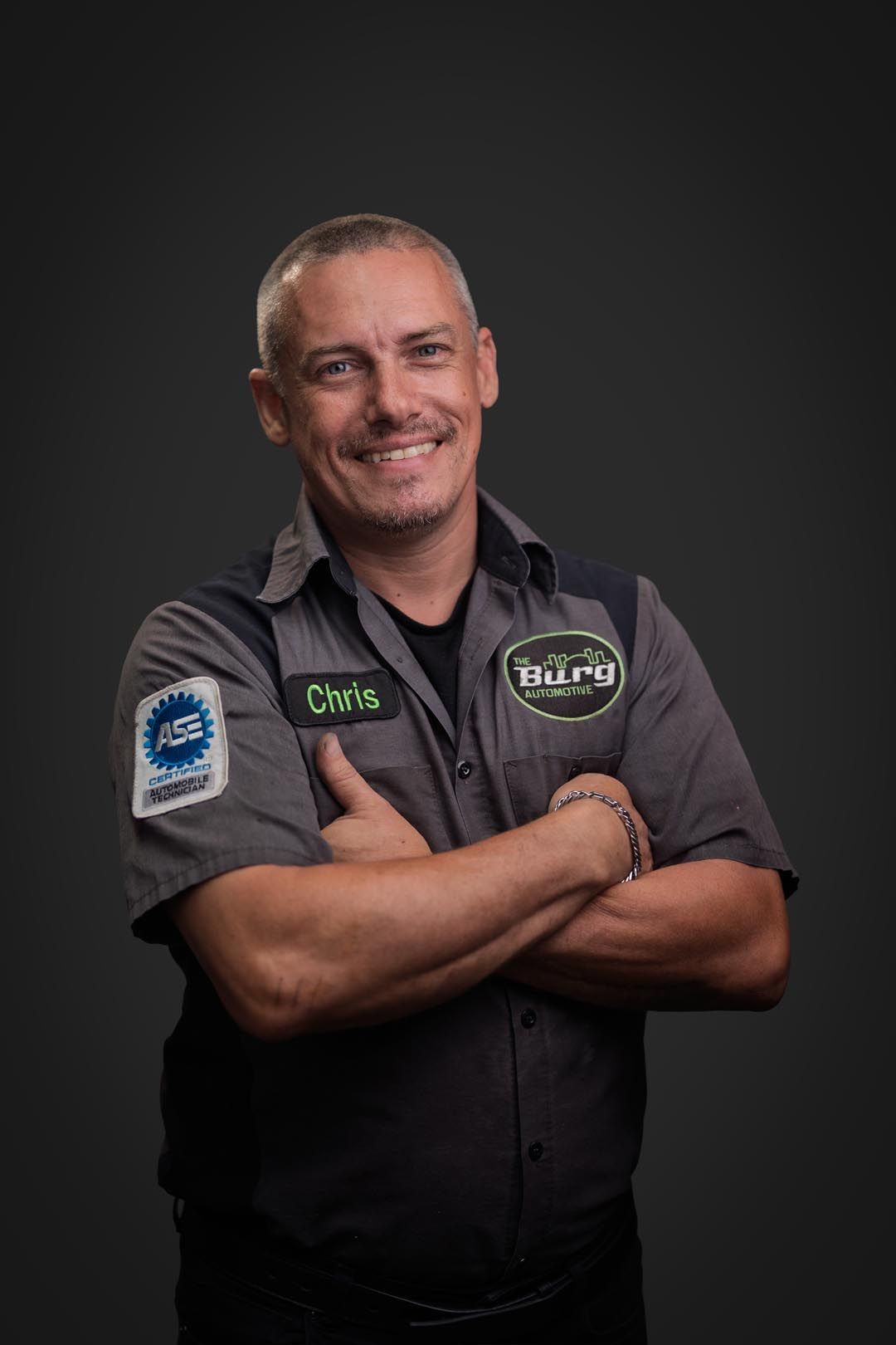 Smiling mechanic in gray work shirt, arms crossed against a dark studio background