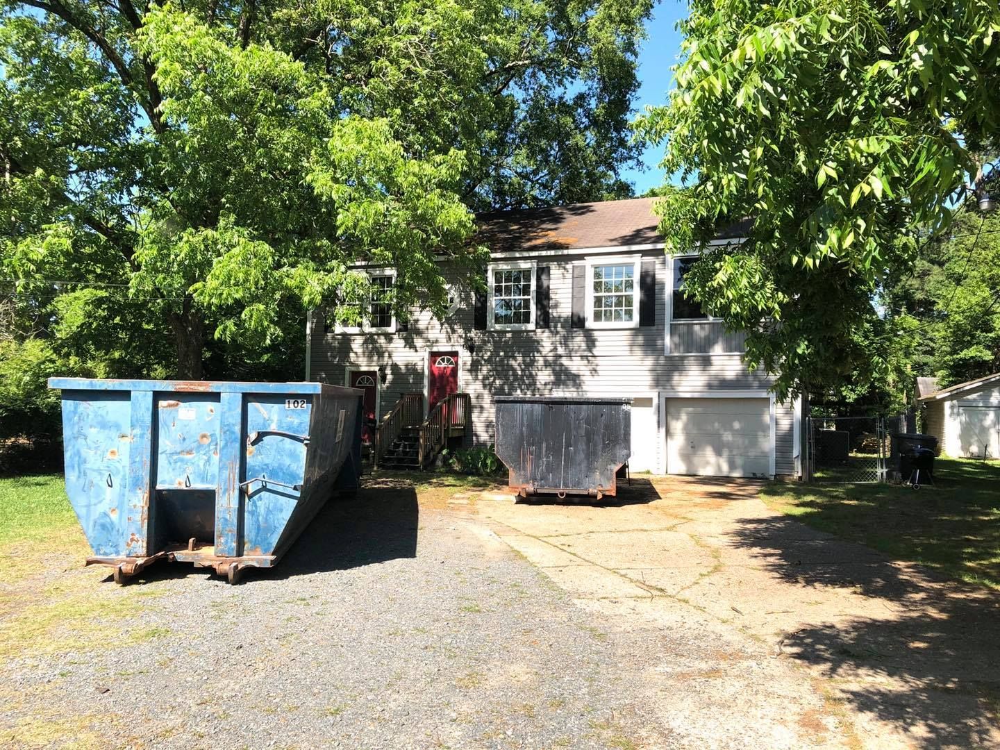 A two-story light-colored house with two large blue construction dumpsters in its gravel driveway.