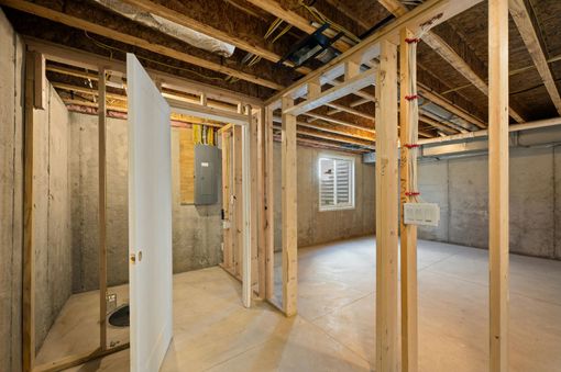 An unfinished basement featuring bare concrete walls, exposed ceiling joists, wooden wall framing, and an electrical panel.