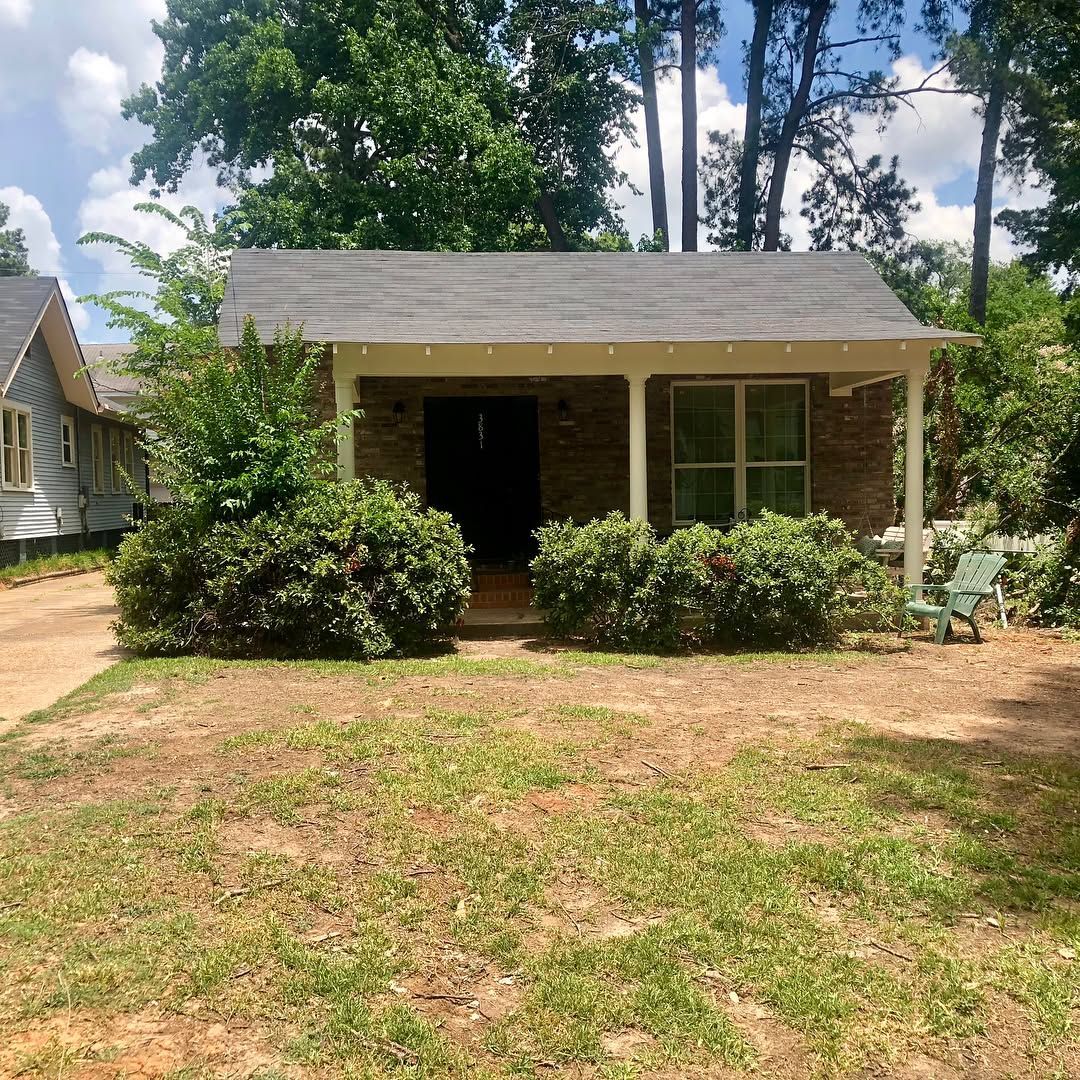 A single-story brick house with a covered front porch and a dark roof, framed by green bushes and trees on a sunny day.