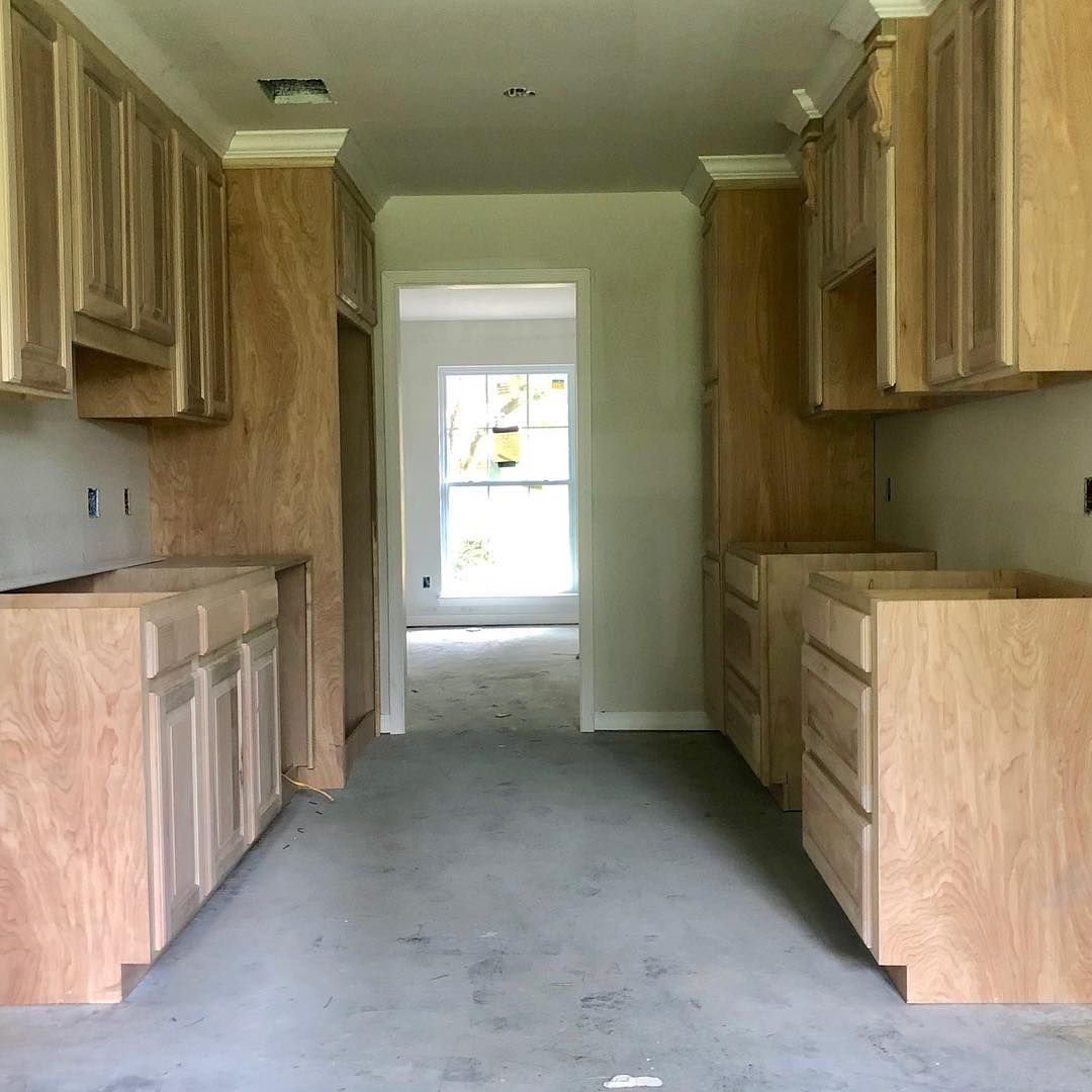 Unfinished kitchen with light wood cabinets installed on both sides of a walkway leading to a doorway with a bright window.