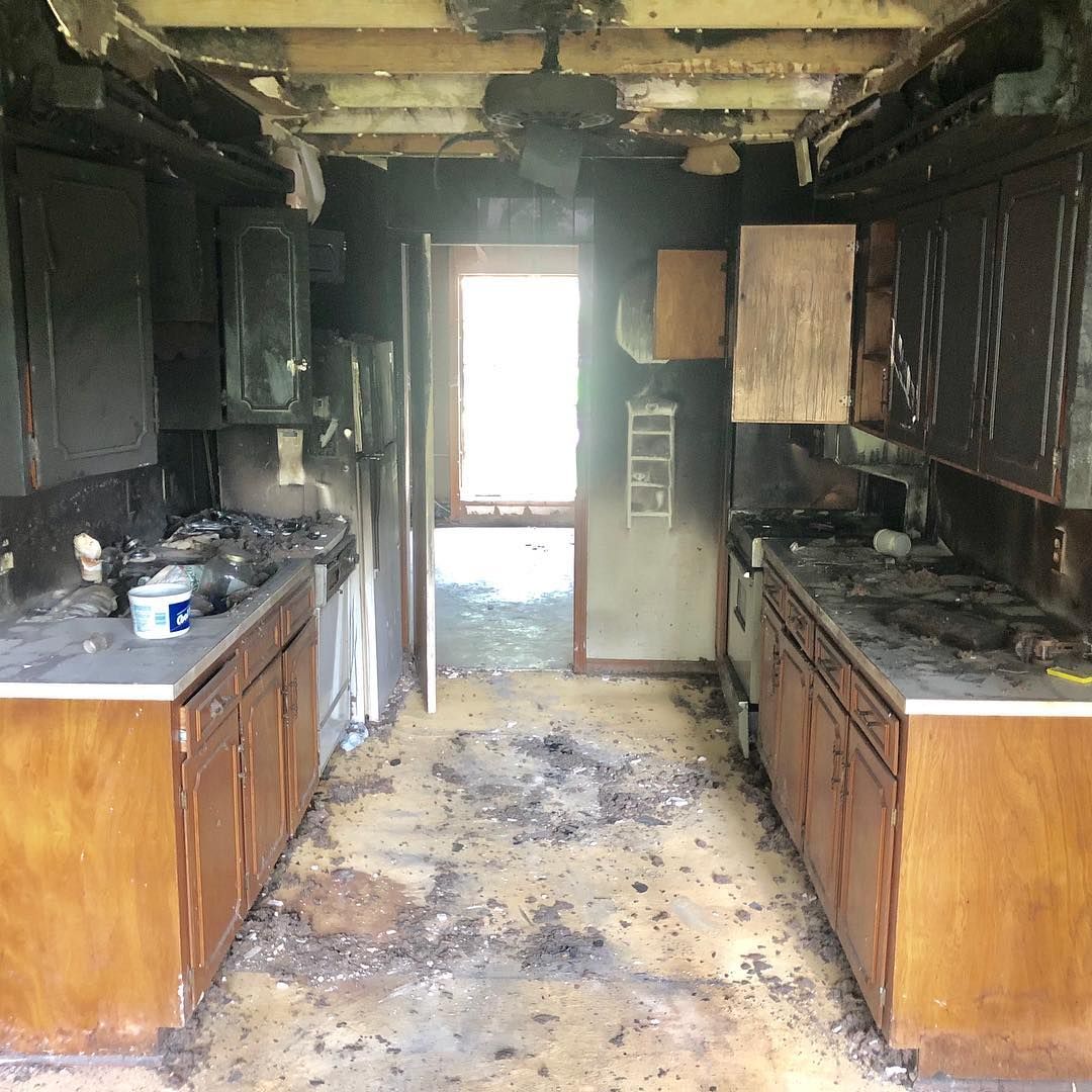 A kitchen interior showing extensive fire and smoke damage, with charred wooden cabinets and debris on the floor.