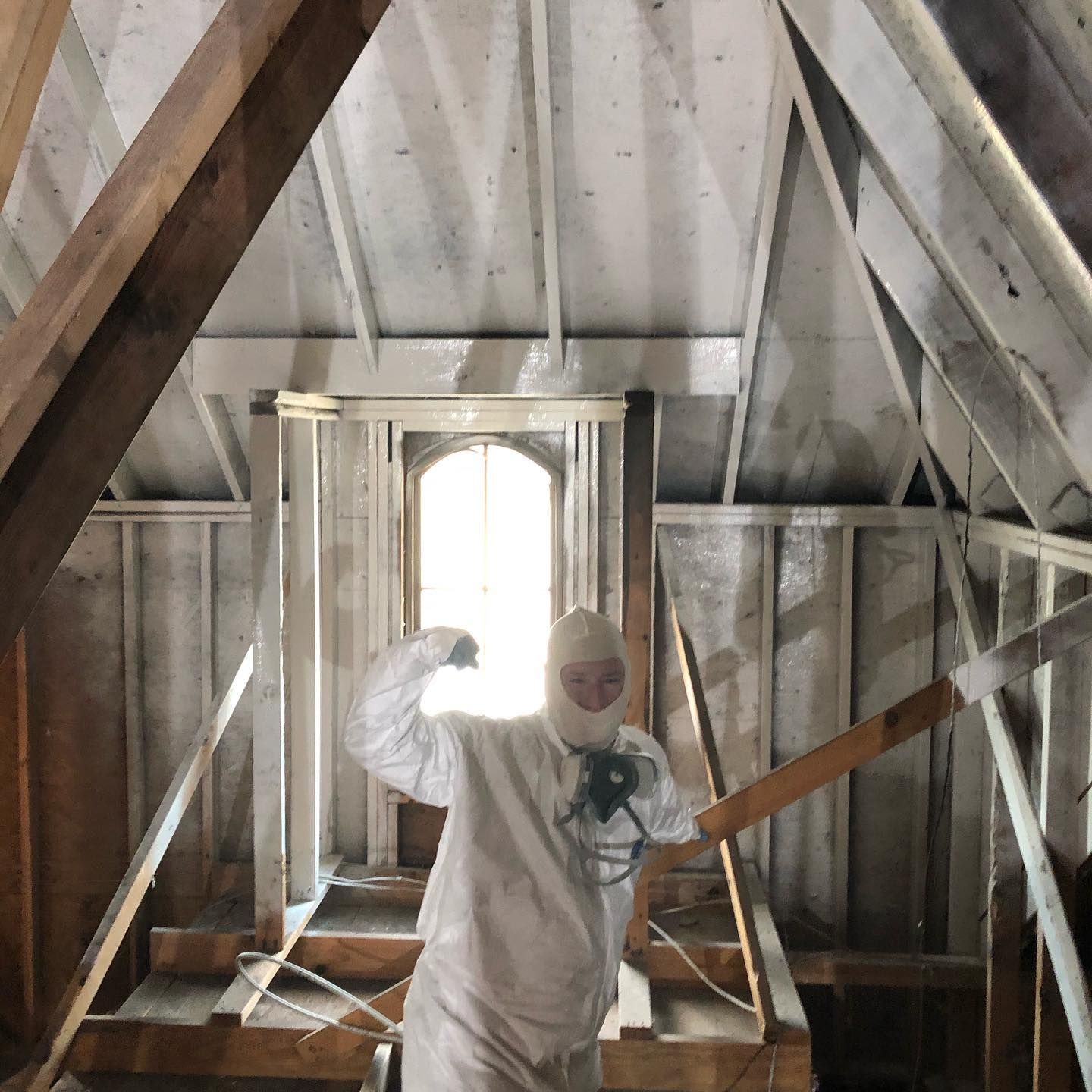 A person in protective white gear and a respirator mask flexes their arm while standing in an unfinished wooden attic.