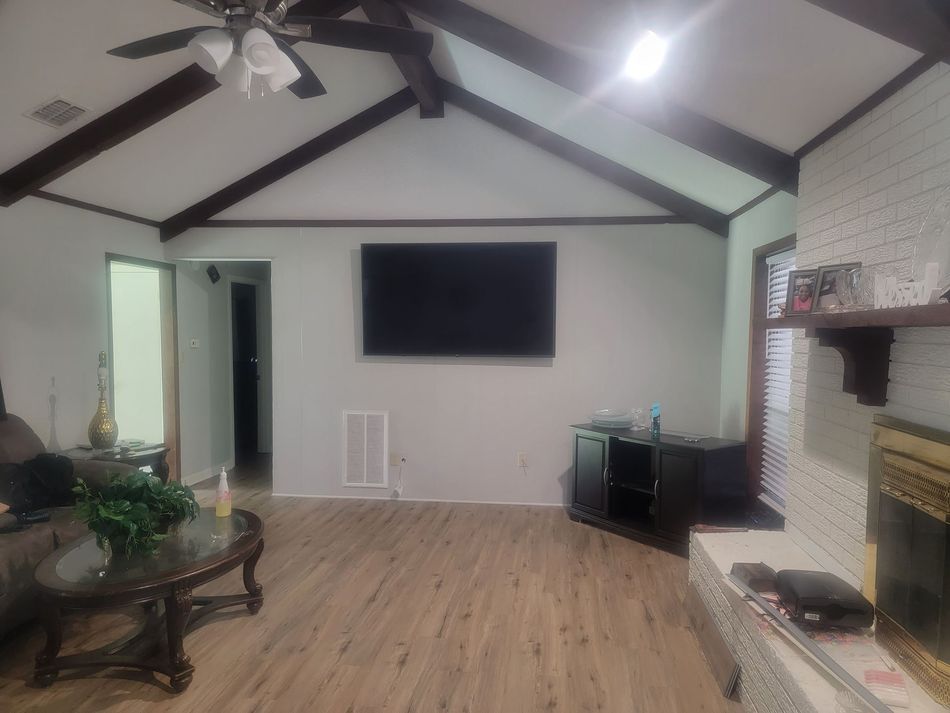 A living room with light wood floors, a vaulted ceiling with dark wooden beams, a mounted TV, and a white brick fireplace.