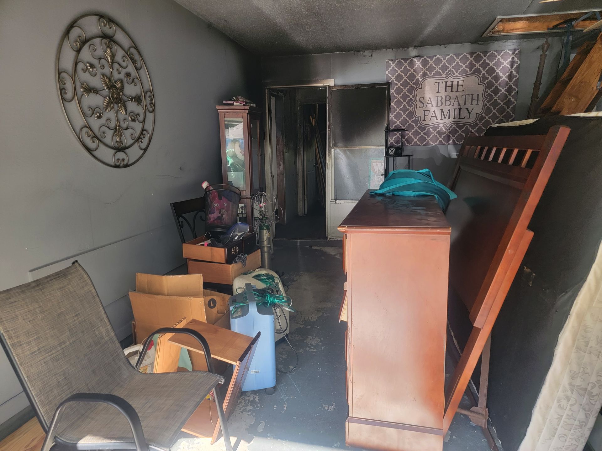 A room with a chair, stacked boxes, and a wooden bed frame, showing signs of fire or smoke damage on the walls and ceiling.