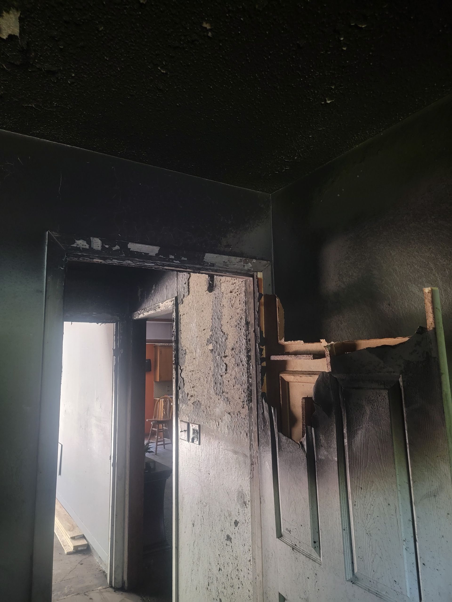 A fire-damaged doorway showing charred, blackened walls and ceiling, looking through into a brightly lit, clean room.