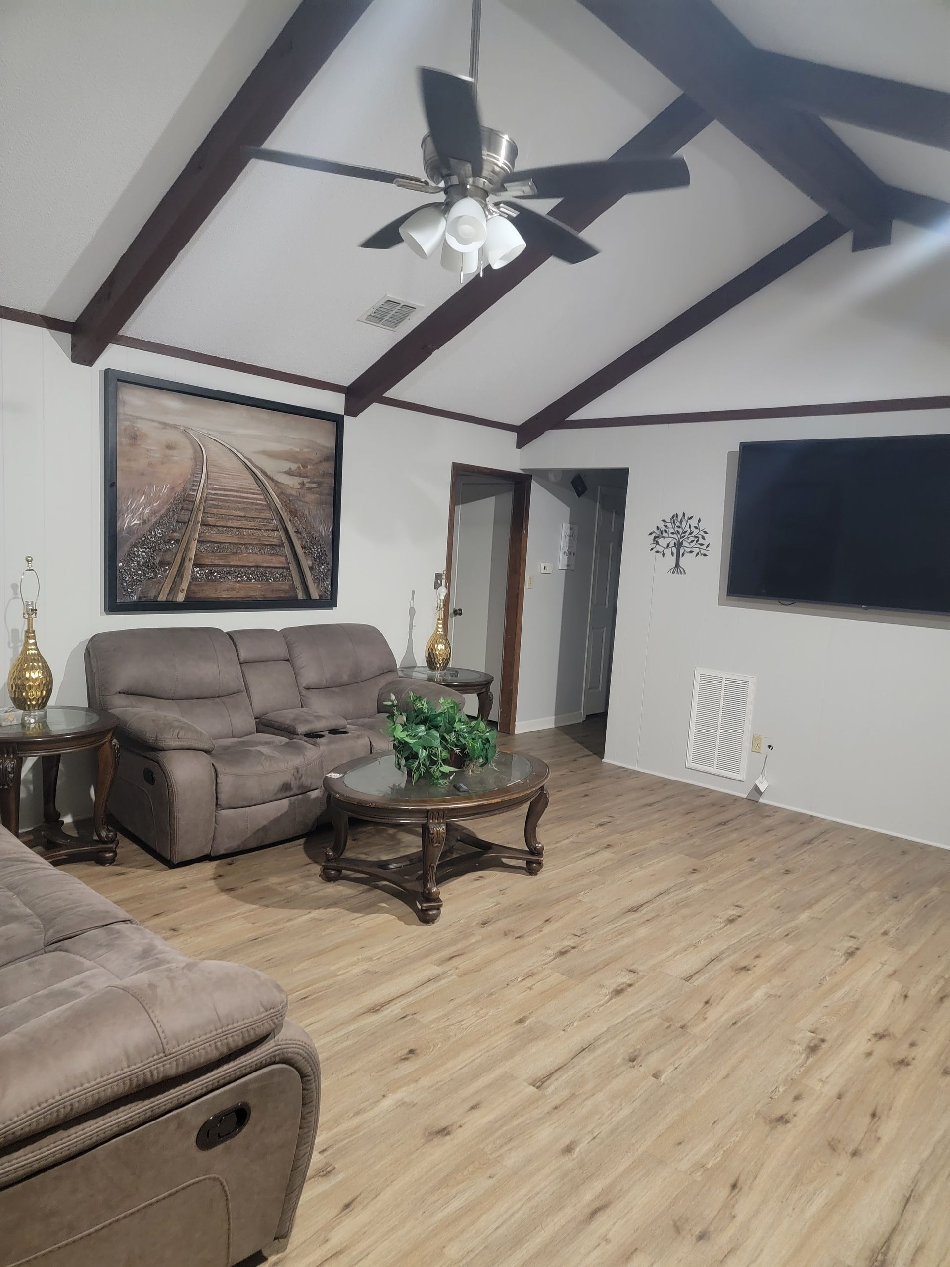 A living room with light wood floors, a vaulted ceiling with dark beams, two brown recliners, and a wall-mounted television.