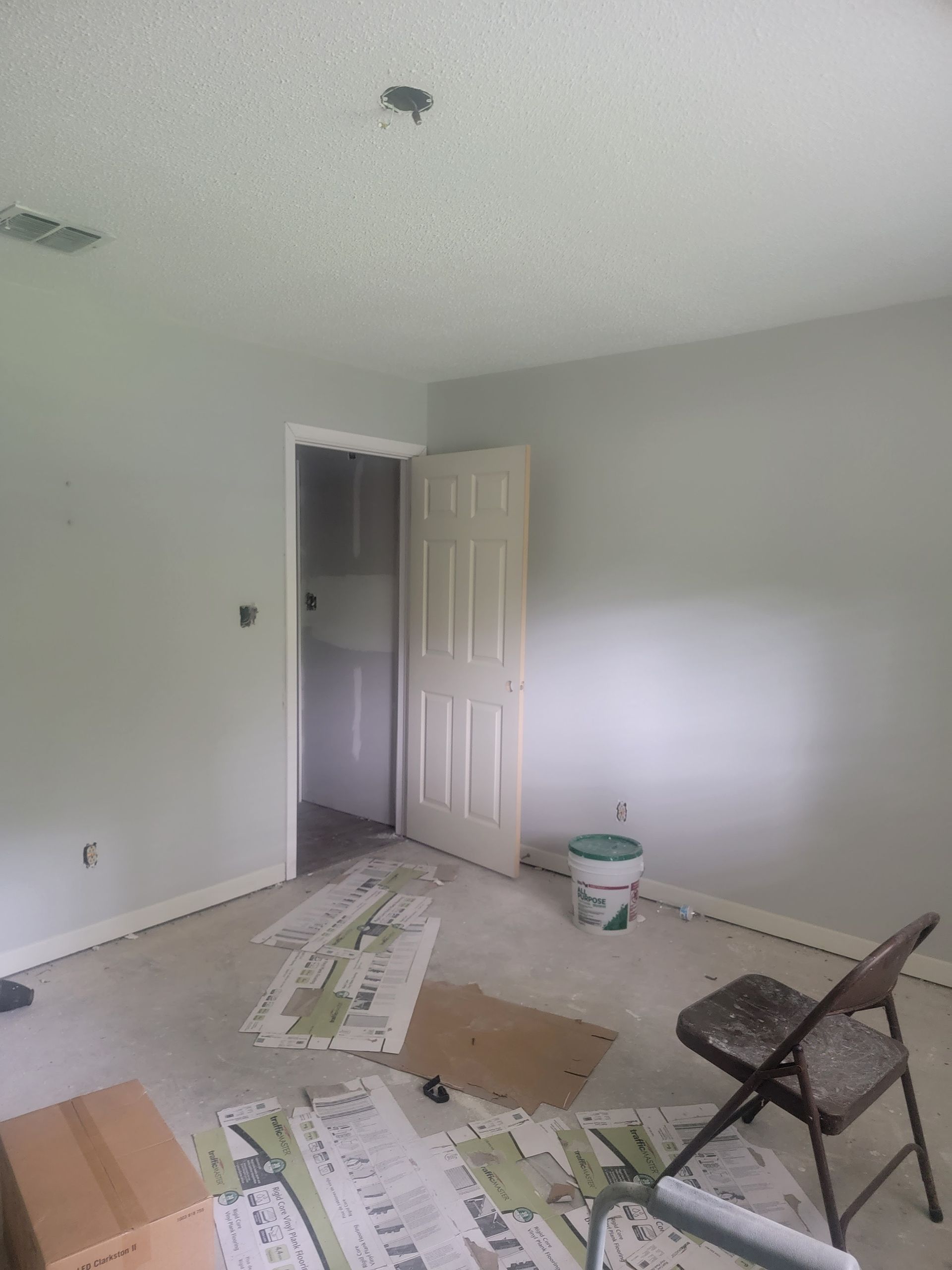 An interior room under renovation with light gray walls, a door, a ceiling fixture, a paint bucket, and debris on the floor.