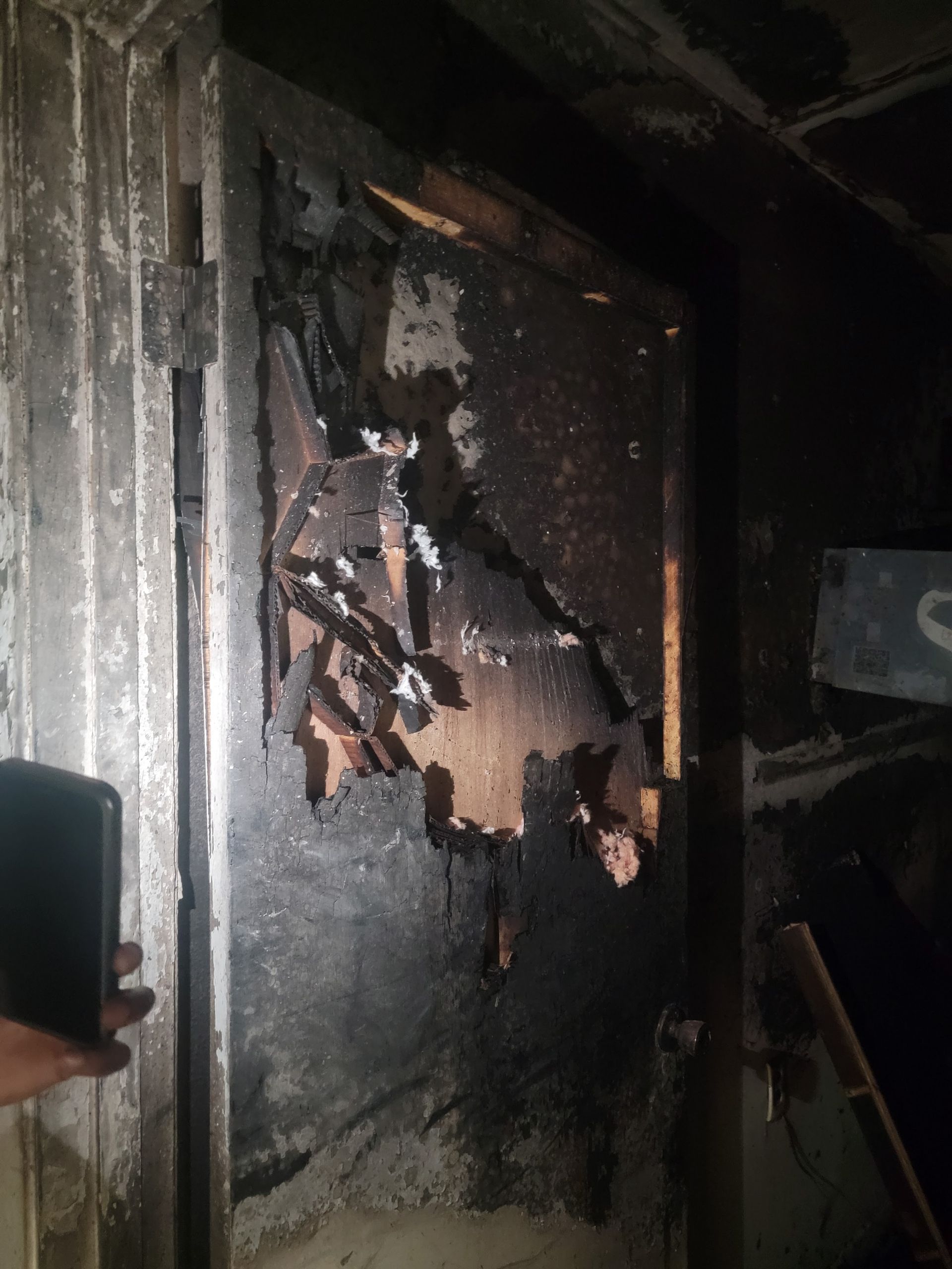 A dark, burnt doorway with charred wooden panels and visible hinges in a dimly lit, damaged room.