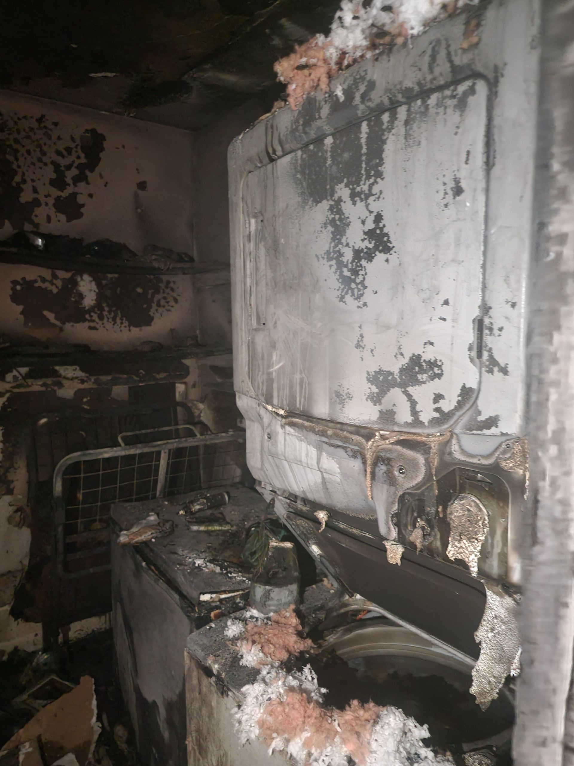 A dark, damaged laundry room with a fire-scorched dryer and washer covered in soot and insulation debris.