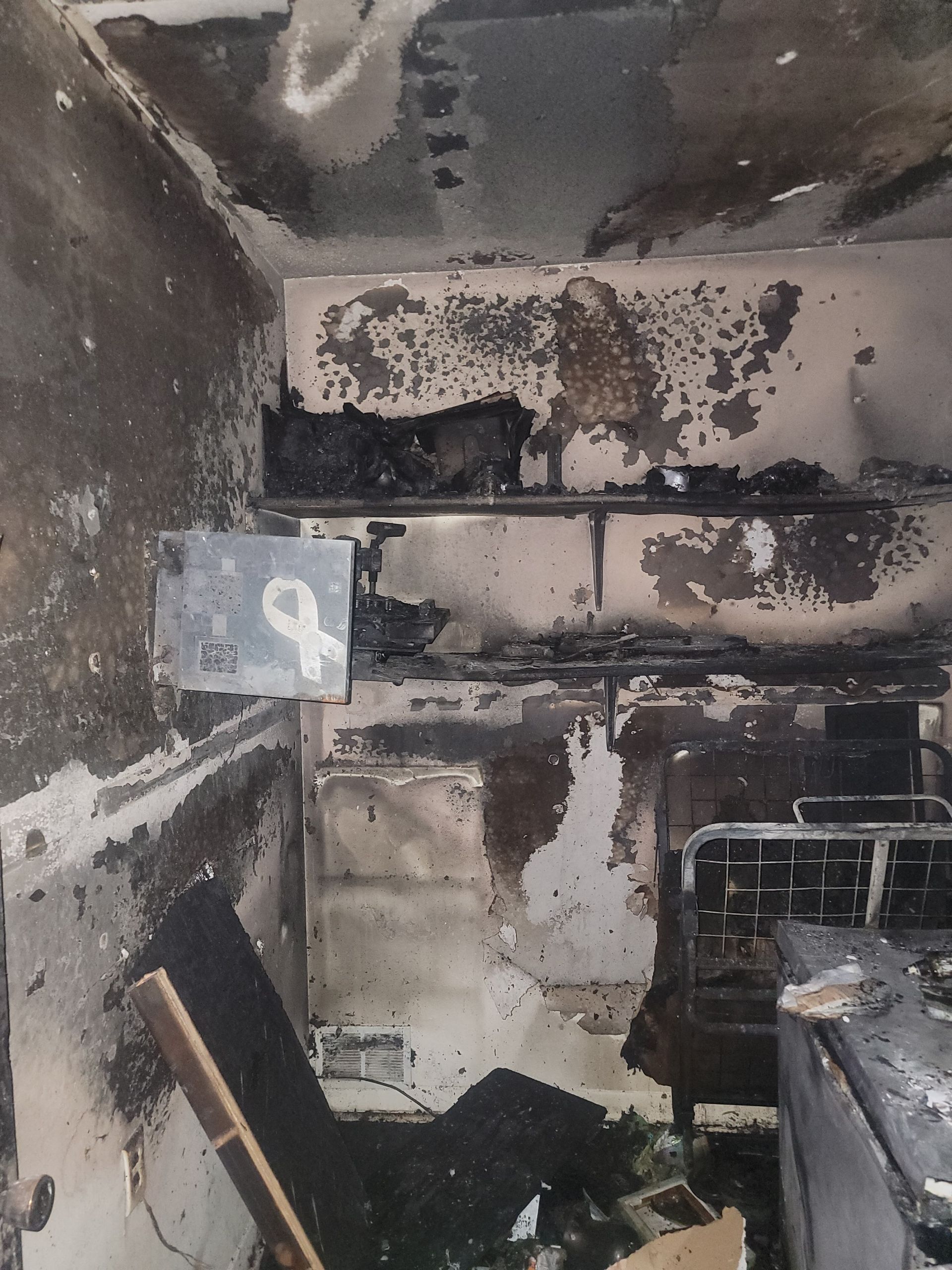 Interior of a room severely damaged by fire, showing charred walls, debris, and a hanging white ribbon sign.
