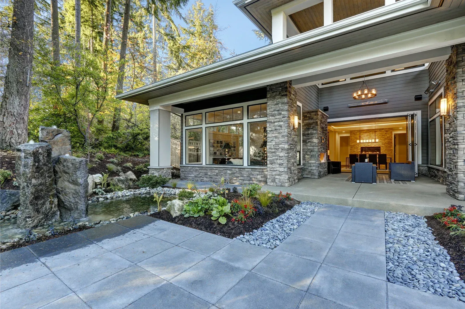 Modern patio with stone walls and large windows leading to an open living area, surrounded by lush trees and garden beds.