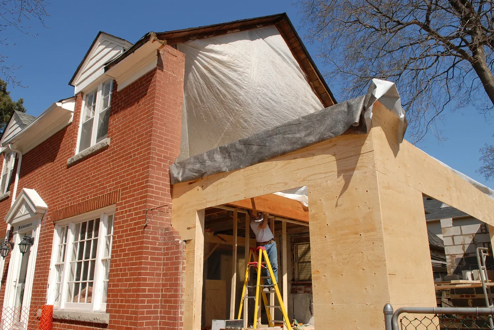 Construction worker on a ladder building a wooden home addition attached to a red brick house under a clear blue sky.