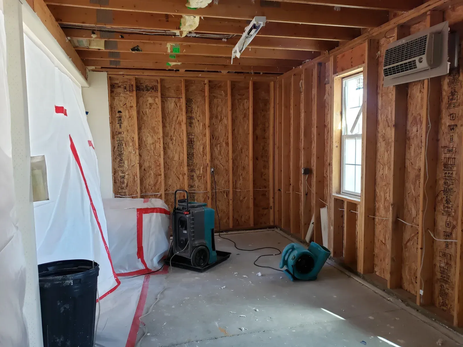 Interior of a room under construction with bare wood studs, a concrete floor, a window, and professional drying equipment.