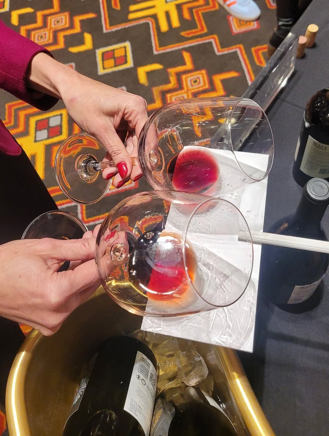 Mujer sosteniendo tres copas de vino con vinos de diferentes colores, junto a botellas de vino.