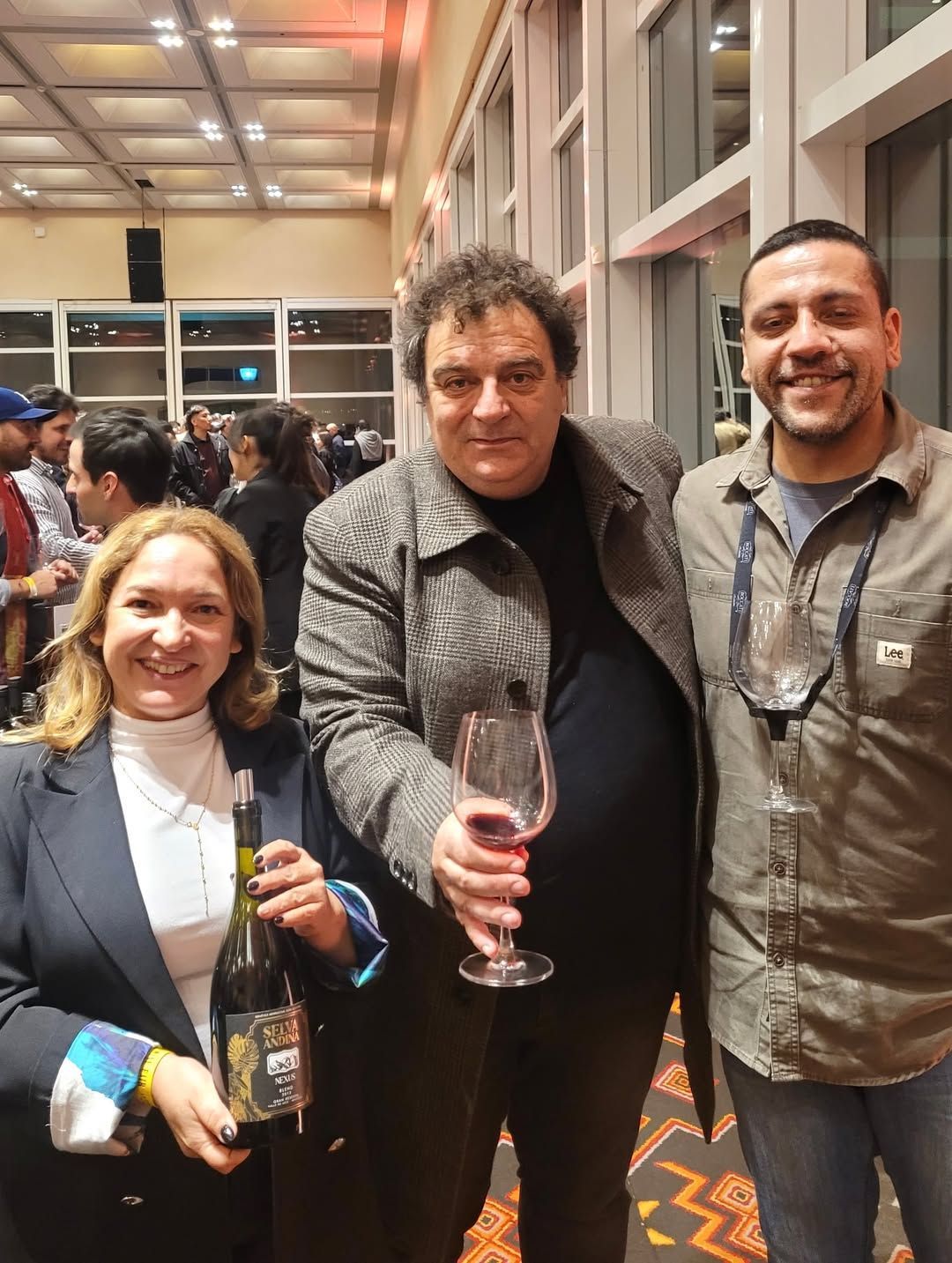 Tres personas en un evento, sosteniendo una botella de vino y una copa; sonriendo en el interior con multitud al fondo.