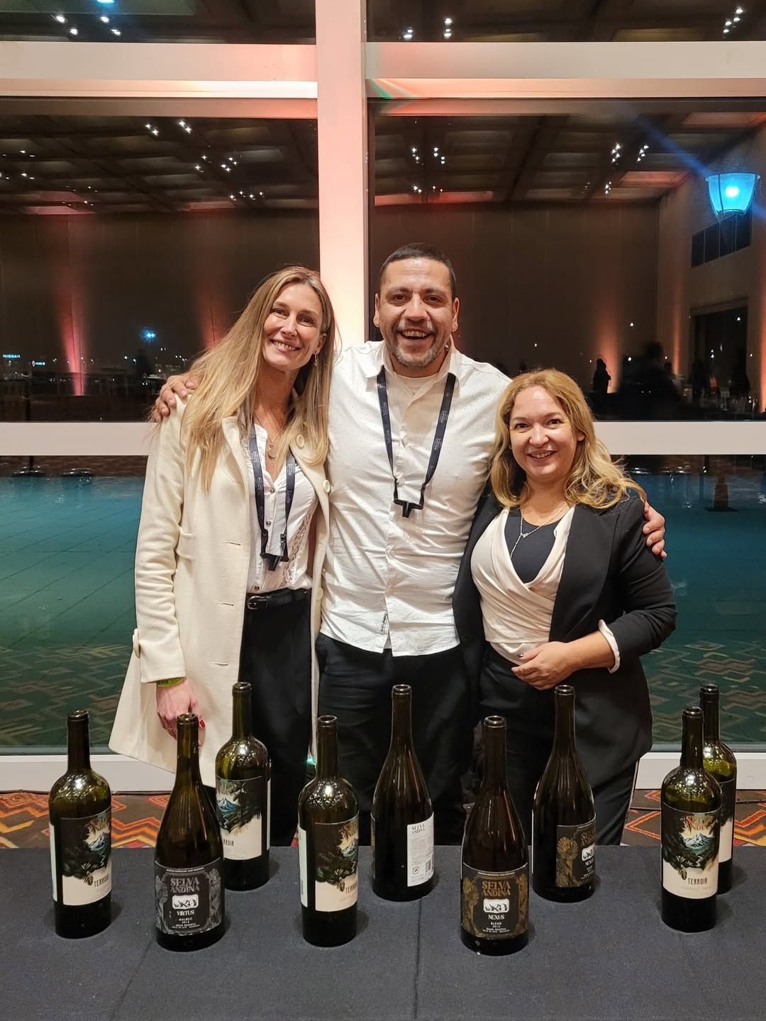 Tres personas sonrientes, posando con botellas de vino sobre una mesa. Ambiente interior, de noche, con agua al fondo.