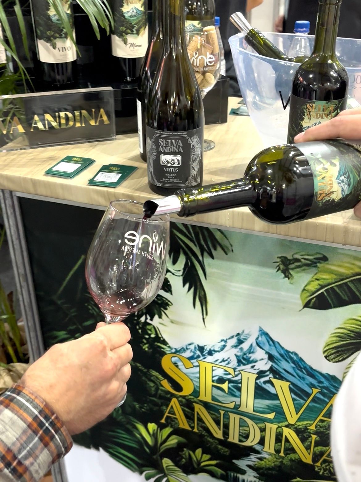 Persona sirviendo vino tinto en una copa en un puesto de cata de vinos; se ven las botellas y el logotipo.