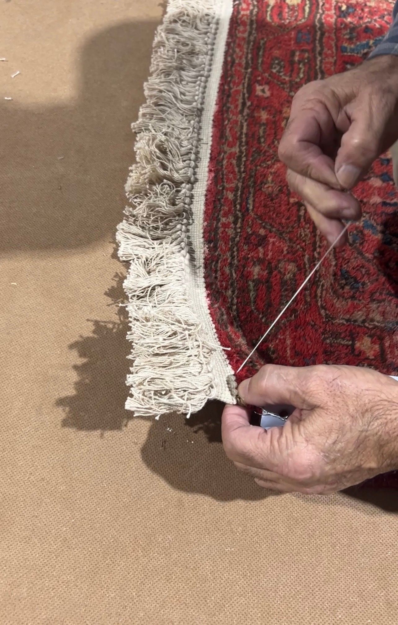 Hands sewing fringe onto a red rug with white fringe, set on a tan surface.