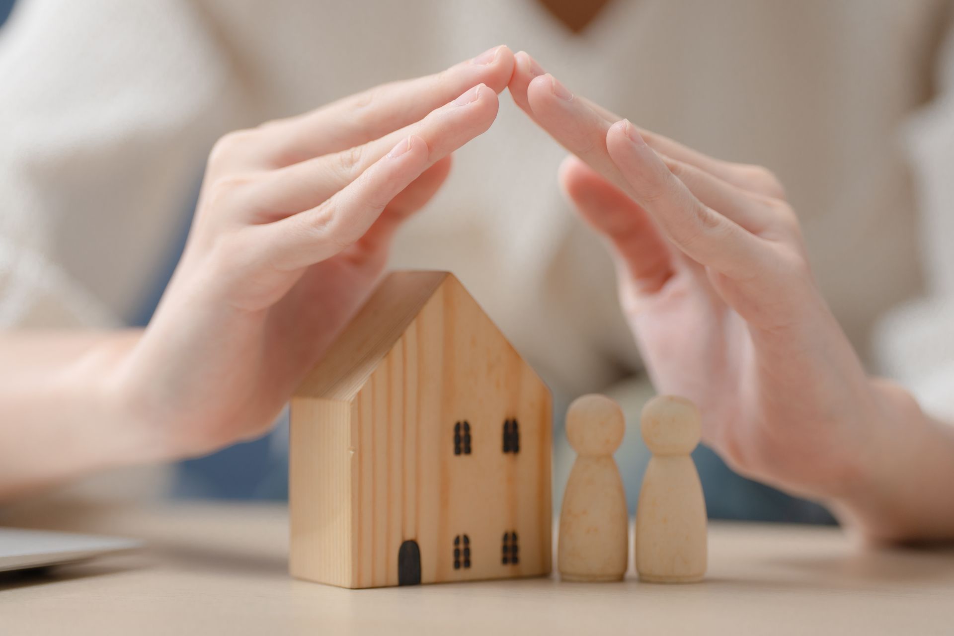 Hands forming a protective roof over a toy house and two wooden figures on a table
