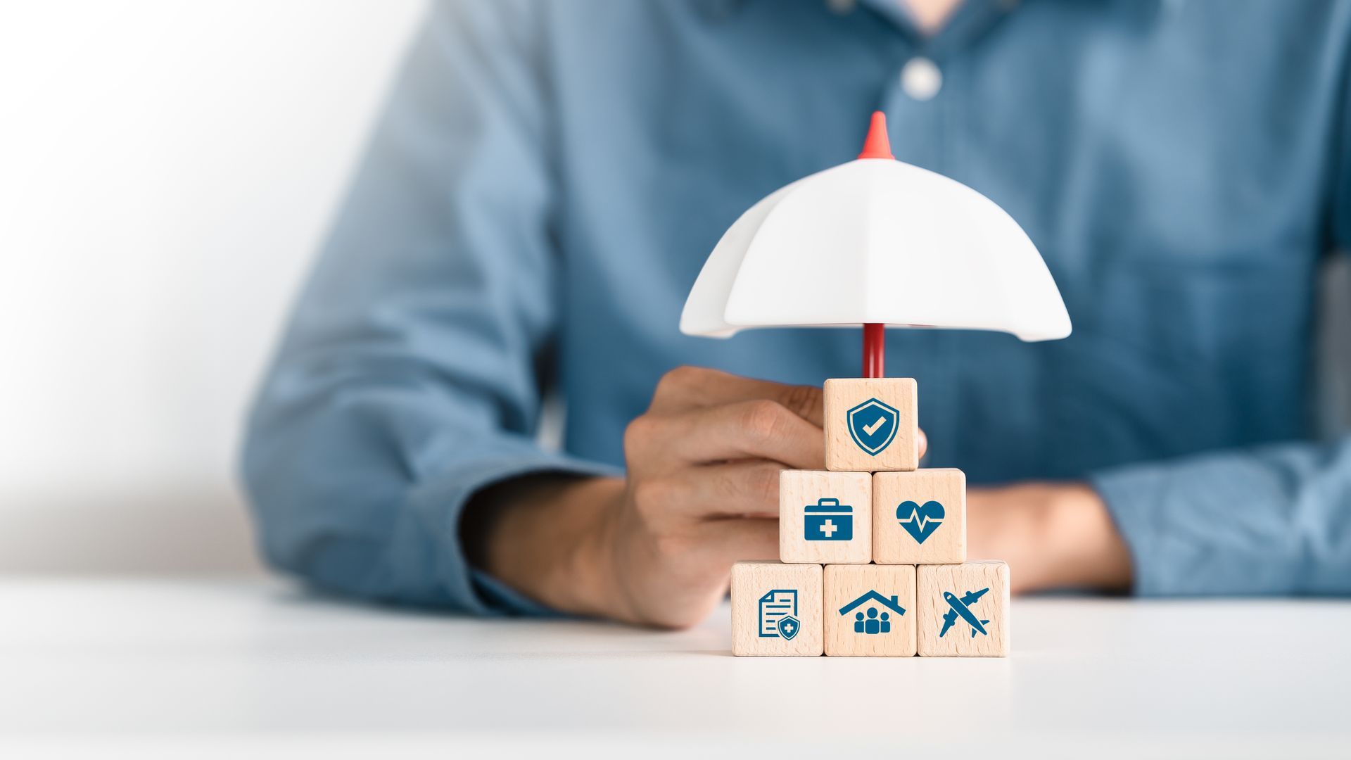 Person holding a small white umbrella over stacked insurance icons on wooden blocks