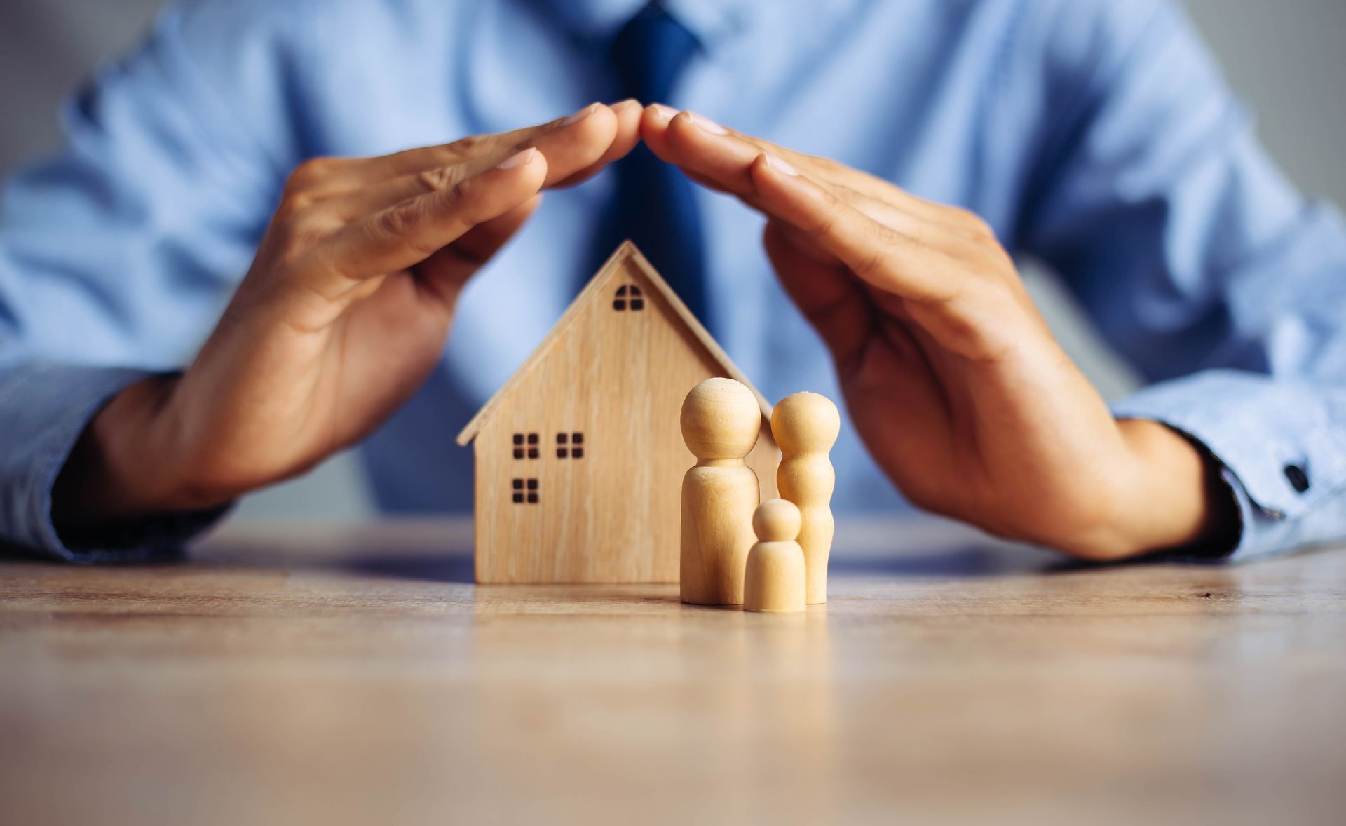 Hands shielding a small wooden house and family figurines on a table, symbolizing home protection