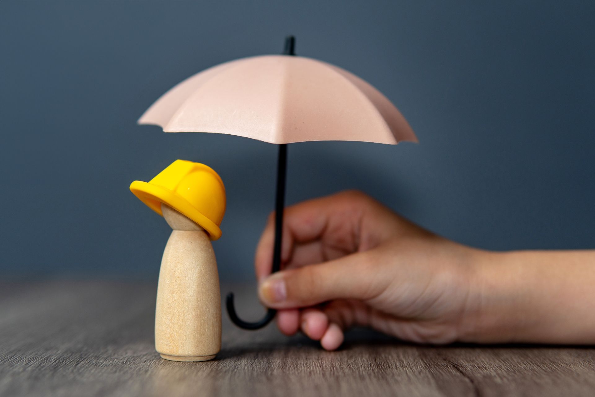 Small figurine in a yellow rain hat beside a hand holding a pale pink umbrella on a wooden table.
