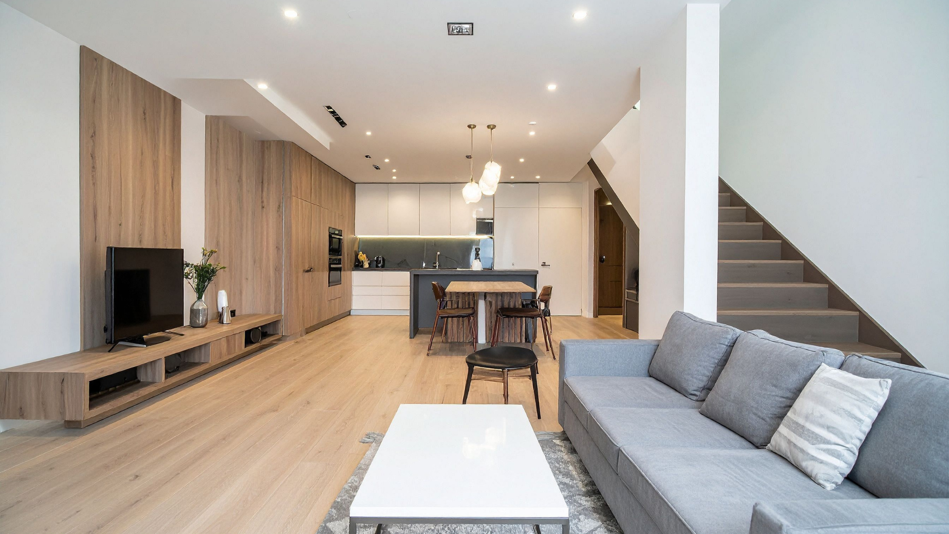 Modern living room with light wood floors, gray sofa, kitchen, and staircase.