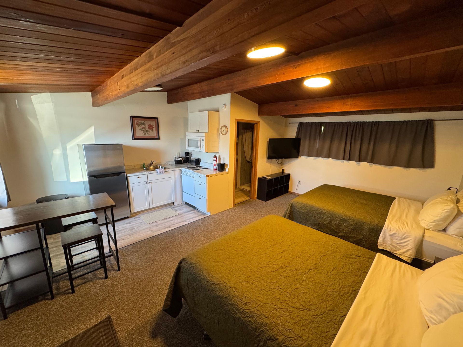 Studio apartment with two beds, a small kitchen, and dining table; brown ceiling beams and dark curtains.